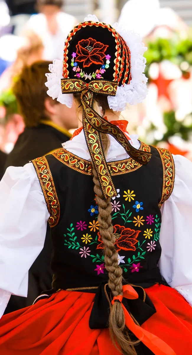 Eine Frau in traditioneller Folklorekleidung ist von hinten zu sehen. Sie trägt ein buntes Kleid mit bestickten Mustern und Bändern.