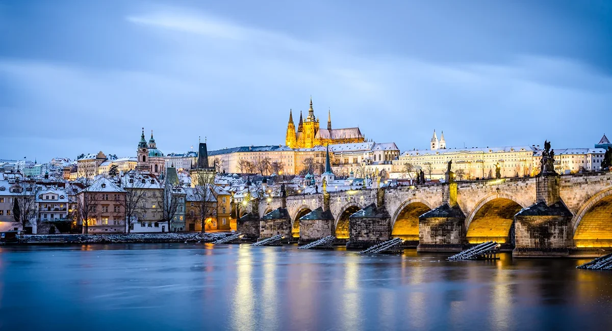 Winterliche Abendstimmung in Prag mit schneebedeckter Brücke und der beleuchteten Burg im Hintergrund.