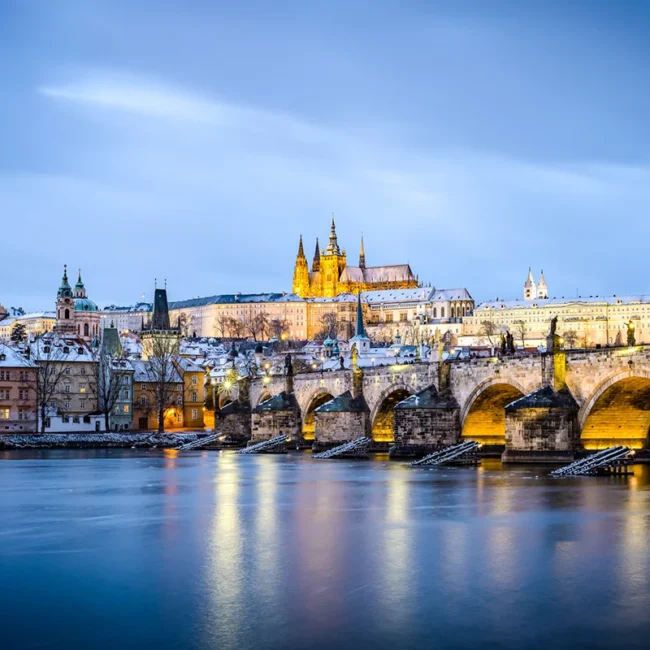 Winterliche Abendstimmung in Prag mit schneebedeckter Brücke und der beleuchteten Burg im Hintergrund.