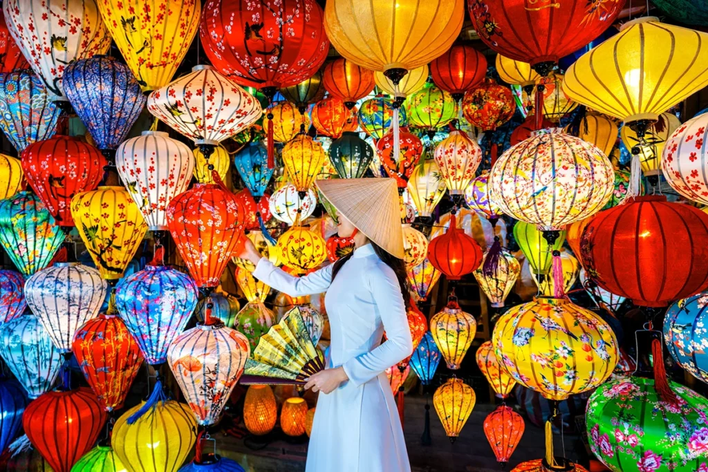 Einheimische Frau im weißen Kleid mit spitzem Hut vor bunten Laternen in Hoi An, Vietnam
