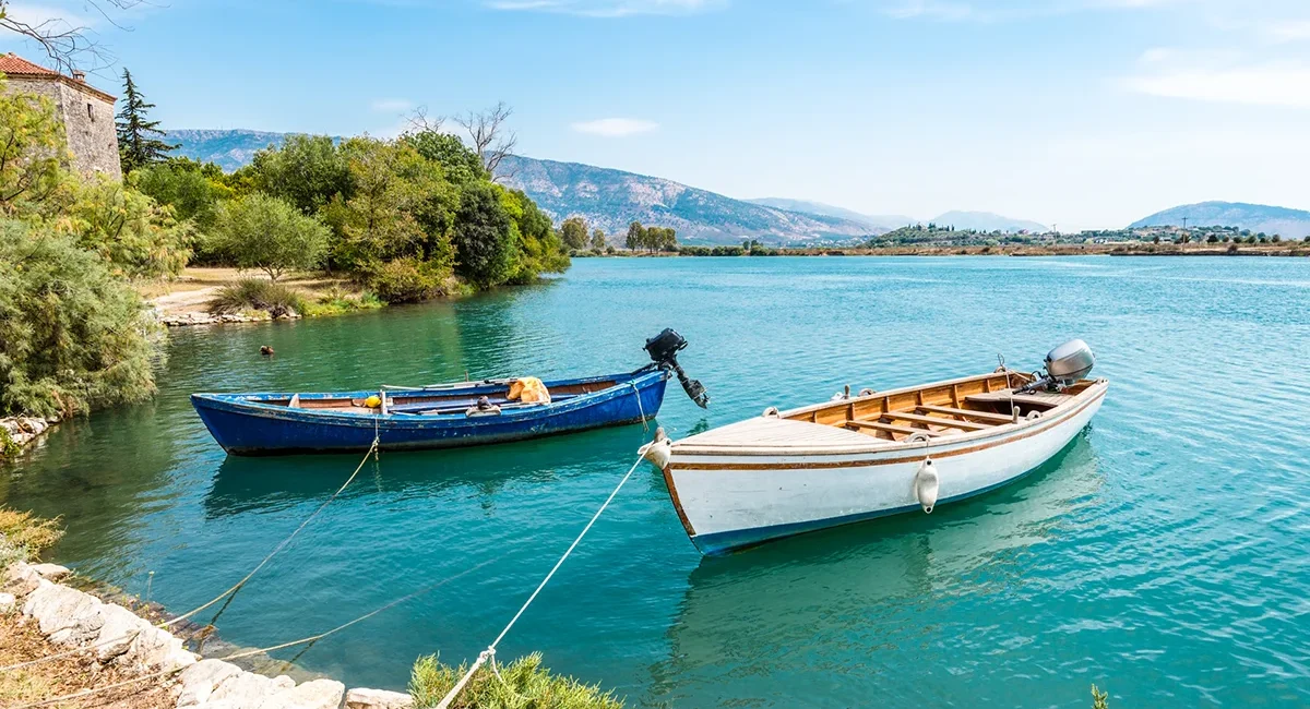 Zwei kleine Motorboote liegen ruhig am bewachsenen Ufer des türkisfarbenen Butrint-Sees in Albanien vor einer malerischen Bergkulisse.