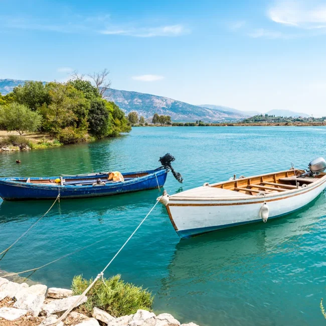 Zwei kleine Motorboote liegen ruhig am bewachsenen Ufer des türkisfarbenen Butrint-Sees in Albanien vor einer malerischen Bergkulisse.