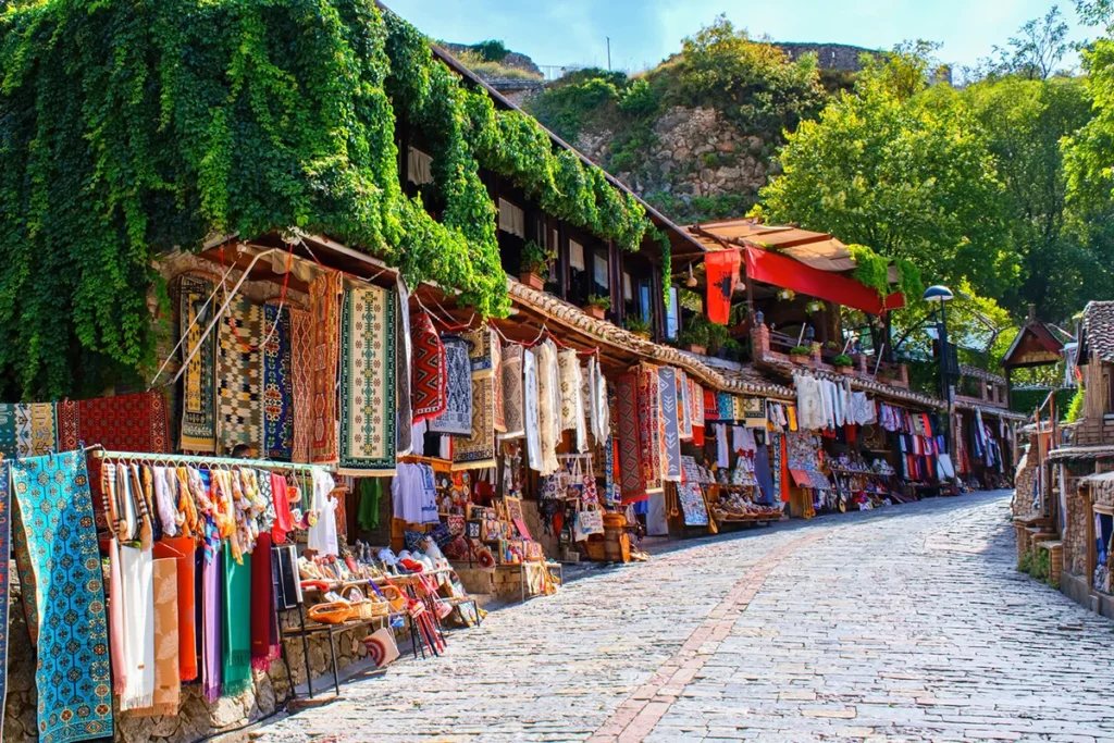 Der farbenfrohe Alte Basar von Kruja in Albanien, eine historische Kopfsteinpflasterstraße mit traditionellen Holzgeschäften, in denen handgefertigte Teppiche und albanisches Kunsthandwerk unter grünen Ranken präsentiert werden.
