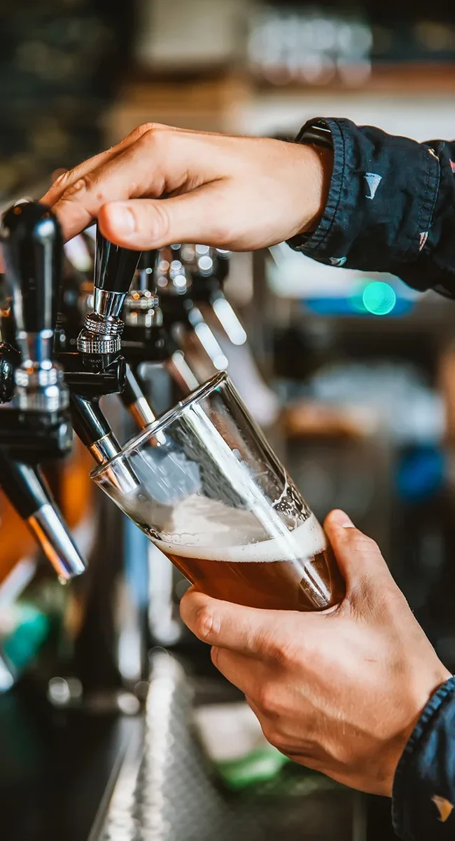 Ein Barkeeper, der an einer professionellen Schankanlage frisches Bier in ein leicht schräg gehaltenes Glas zapft.