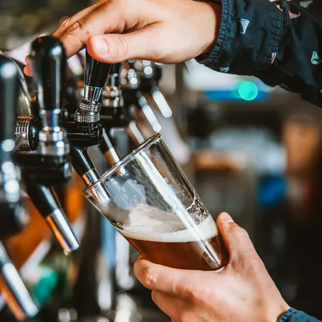 Ein Barkeeper, der an einer professionellen Schankanlage frisches Bier in ein leicht schräg gehaltenes Glas zapft.