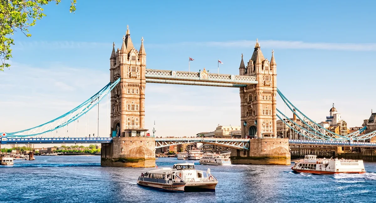 Die berühmte Londoner Tower Bridge in England bei strahlendem Sonnenschein über der Themse, während mehrere Ausflugsschiffe auf dem Wasser unterwegs sind.