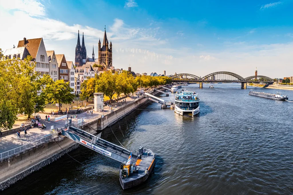 Panoramablick auf den Rhein in Köln mit einem Schiffsanleger am Ufer und der Stadt im Hintergrund.