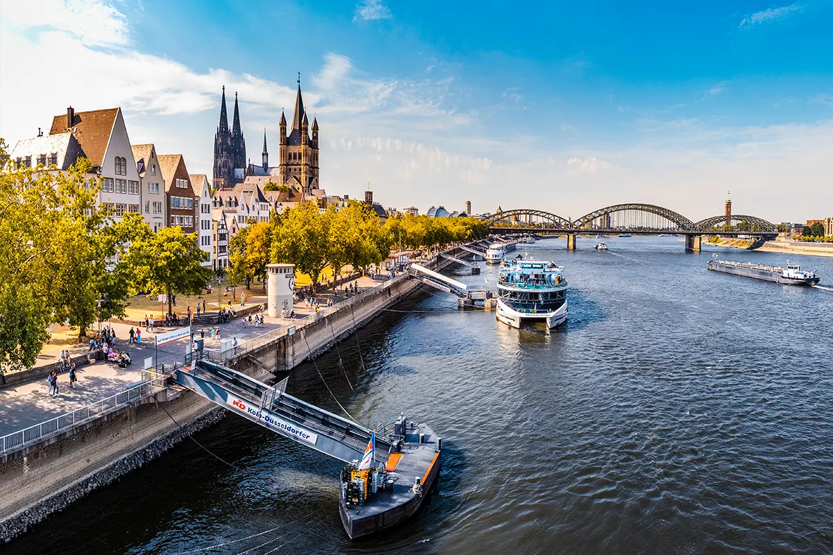Panoramablick auf den Rhein in Köln mit einem Schiffsanleger am Ufer und der Stadt im Hintergrund.