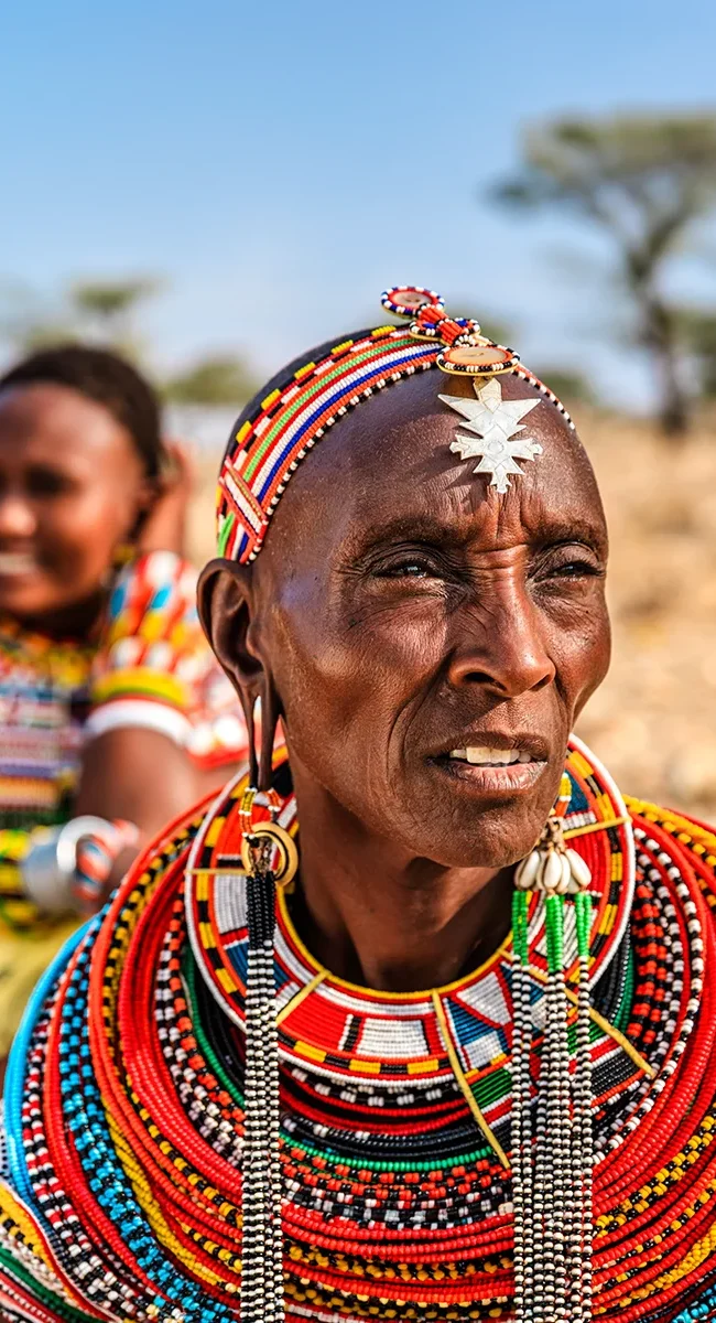 Eine ältere Frau der Massai in ihrer traditionellen, farbenprächtigen Kleidung und mit aufwendigem Perlenschmuck vor einer sonnigen, trockenen Landschaft in Kenia.