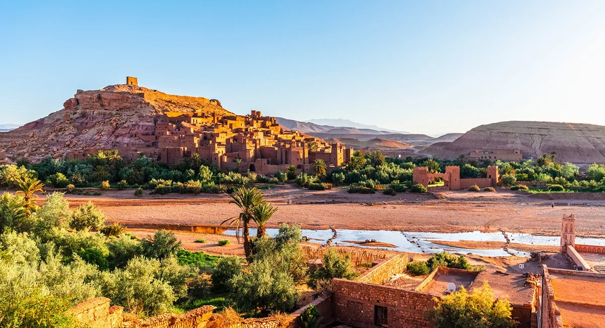 Den Ksar Aït-Ben-Haddou, eine historische Lehmsiedlung in Marokko, die vor einer kargen Wüstenlandschaft malerisch an einem Felshang über einem Flussbett liegt.