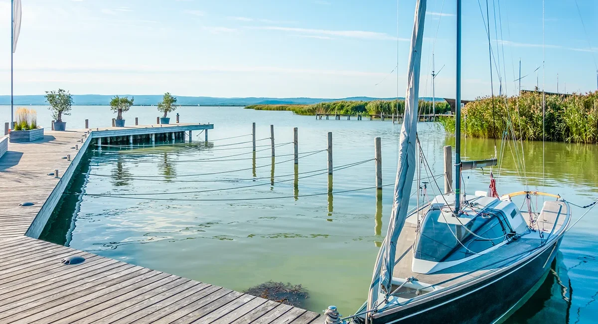 Ein Segelboot liegt an einem Holzsteg in einem modernen Hafen am Neusiedler See in Ungarn bei strahlendem Sonnenschein und angenehmer Atmosphäre.