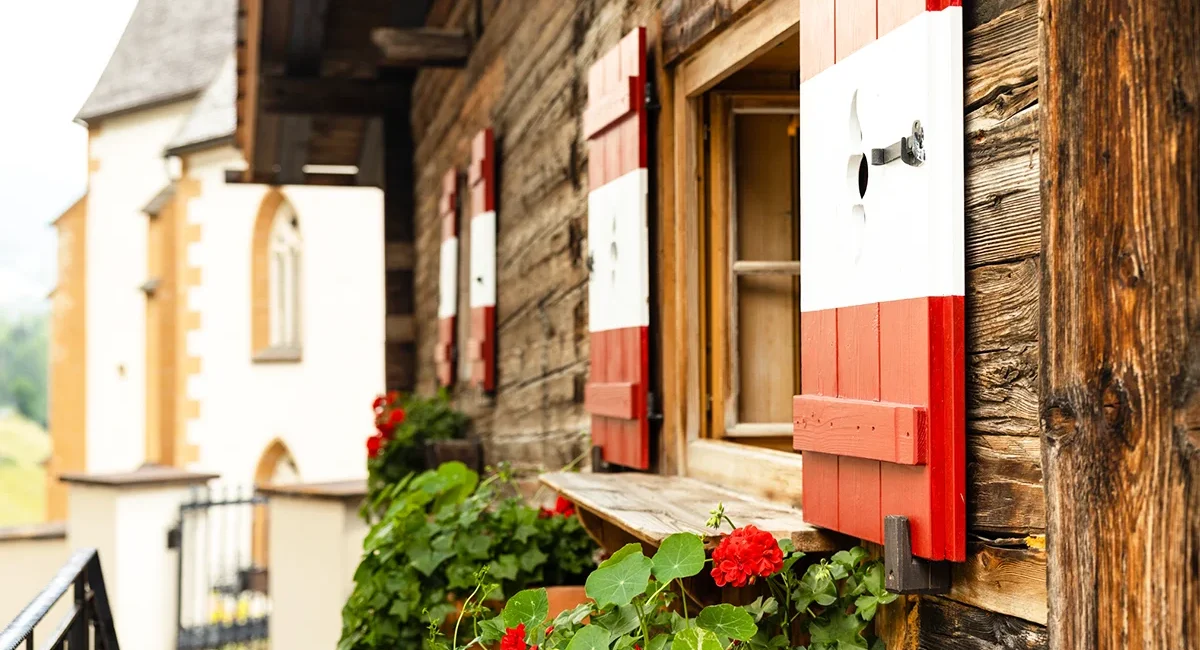Ein rustikales Bauernhaus in Vorarlberg mit rot-weißen Fensterläden, Geranien und einer Kirche im Hintergrund.