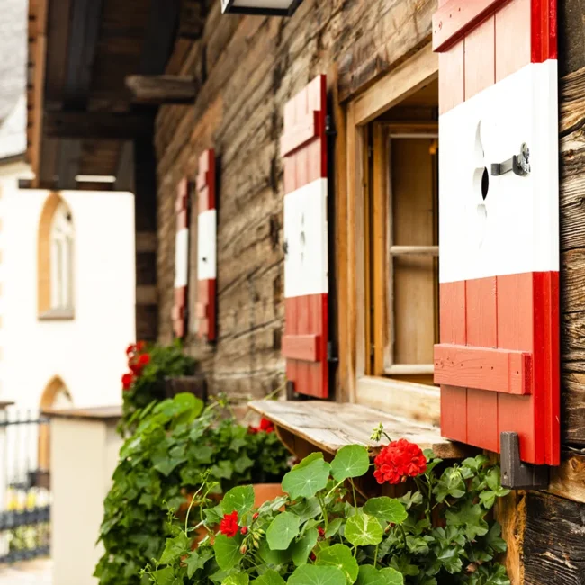 Ein rustikales Bauernhaus in Vorarlberg mit rot-weißen Fensterläden, Geranien und einer Kirche im Hintergrund.