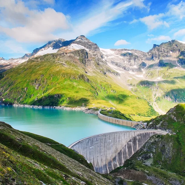 Eine Gebirgslandschaft mit zwei großen Staumauern und dem Wasserkraftwerk der Illwerke in Vorarlberg, die einen türkisblauen See inmitten grüner Hänge und schneebedeckter Gipfel einrahmen.