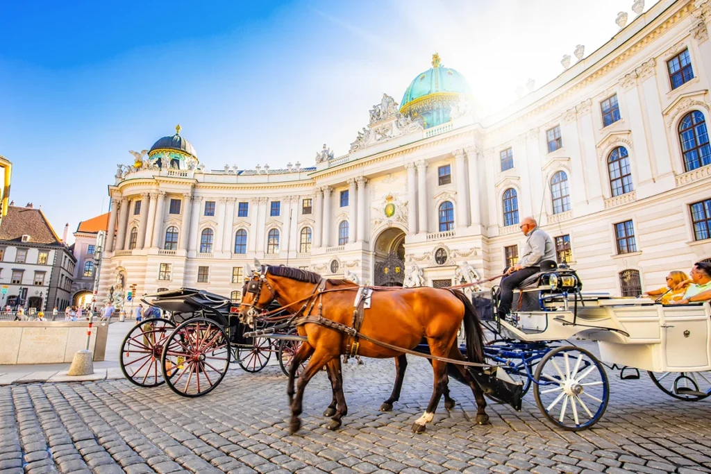 Zwei traditionelle Fiaker-Pferdekutschen auf dem gepflasterten Michaelerplatz vor der prachtvollen, neobarocken Fassade der Wiener Hofburg unter strahlend blauem Himmel.