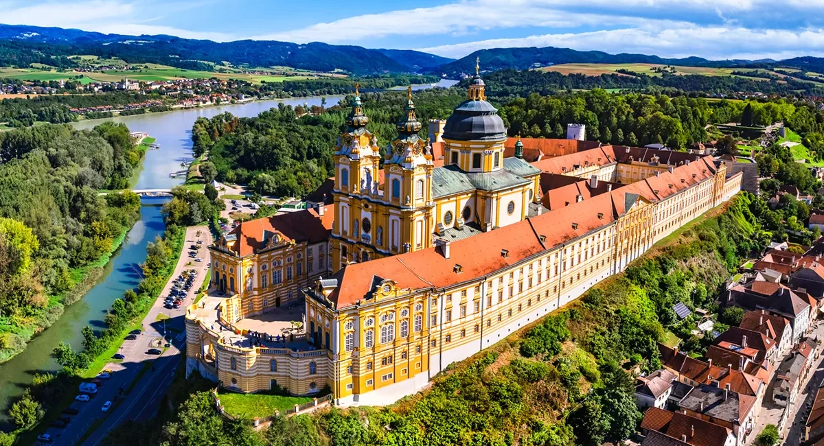 Das prachtvolle, gelb-weiße Stift Melk in der Wachau, das auf einem bewaldeten Hügel direkt über einer Schleife der Donau thront und von einer hügeligen, grünen Landschaft umgeben ist.